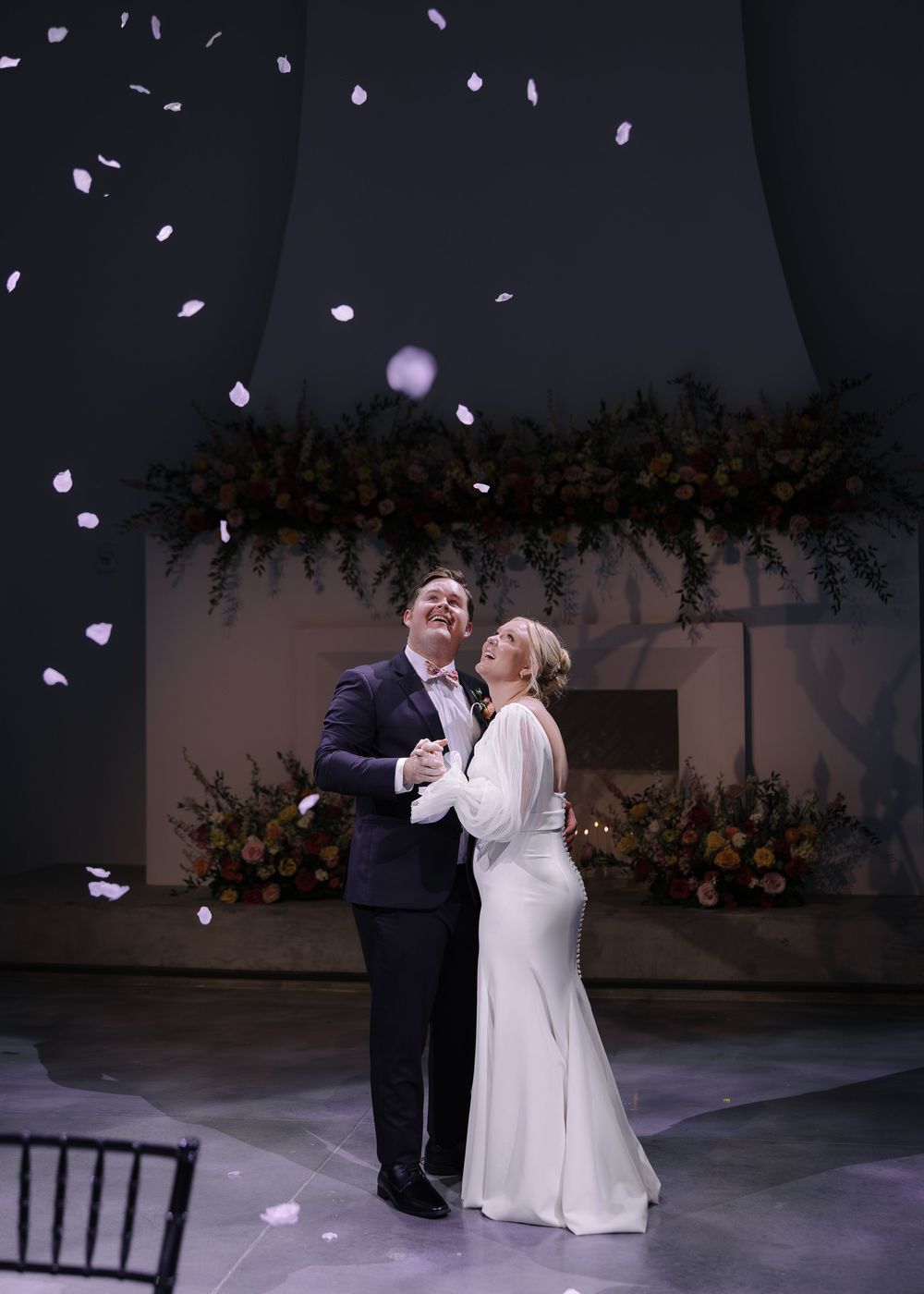 A couple shares their first dance as flower petals fall from above during an elegant indoor wedding ceremony.