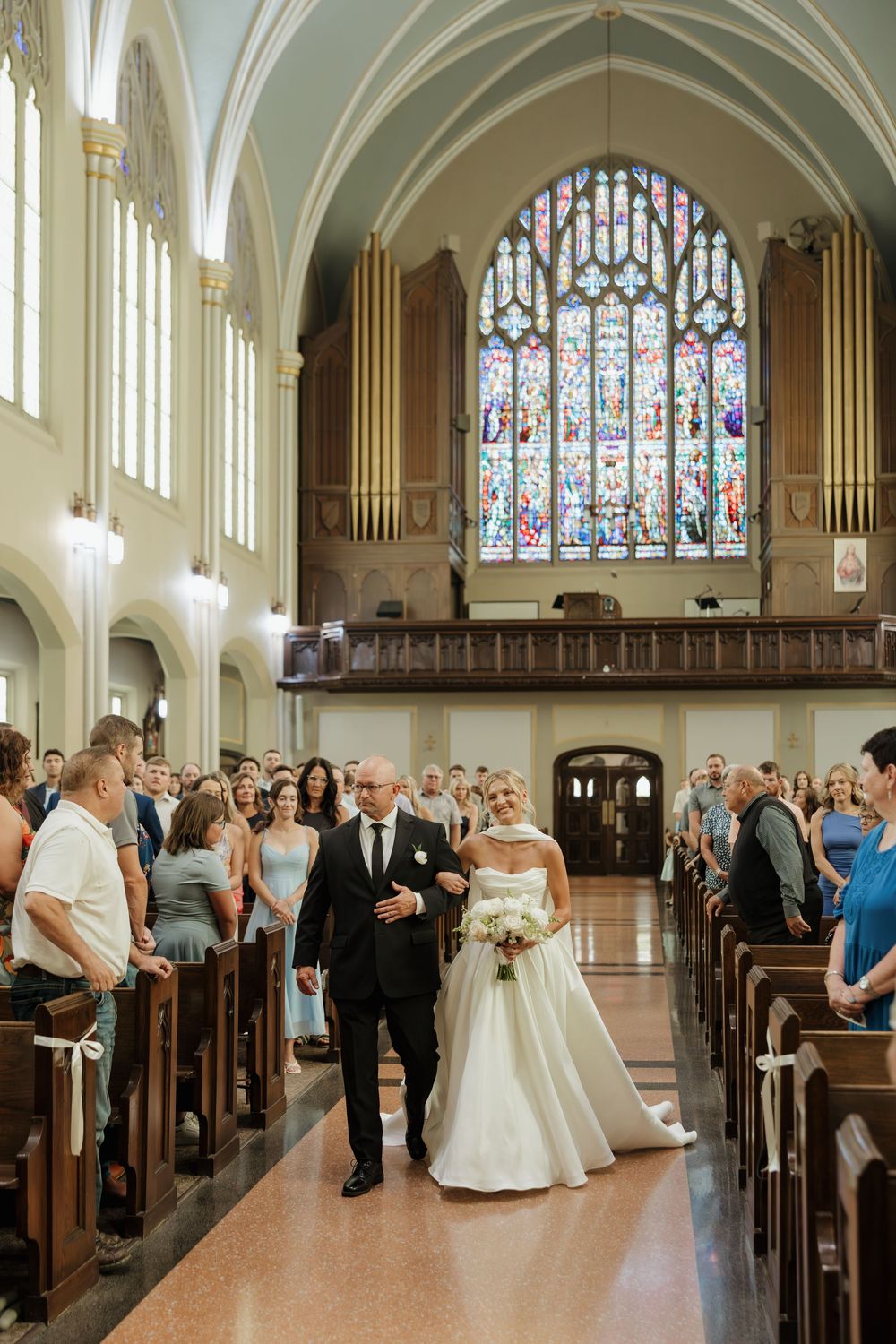 A panoramic view of a traditional church wedding ceremony with guests seated in wooden pews beneath stained glass windows.