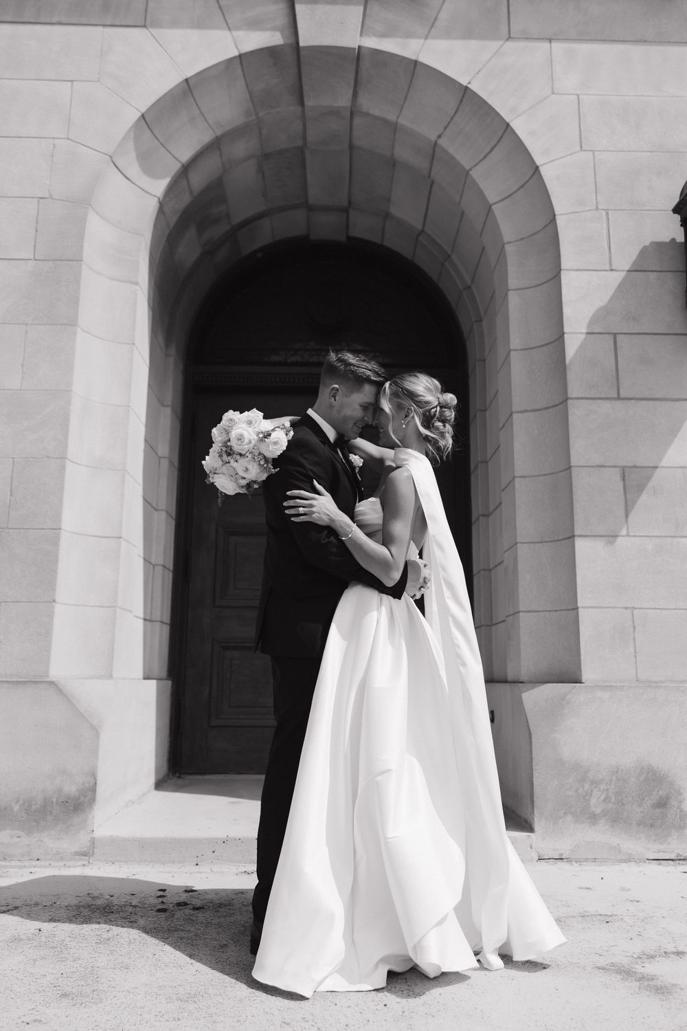 Black and white wedding photo showing a couple embracing in an arched stone doorway with the bride holding white flowers.