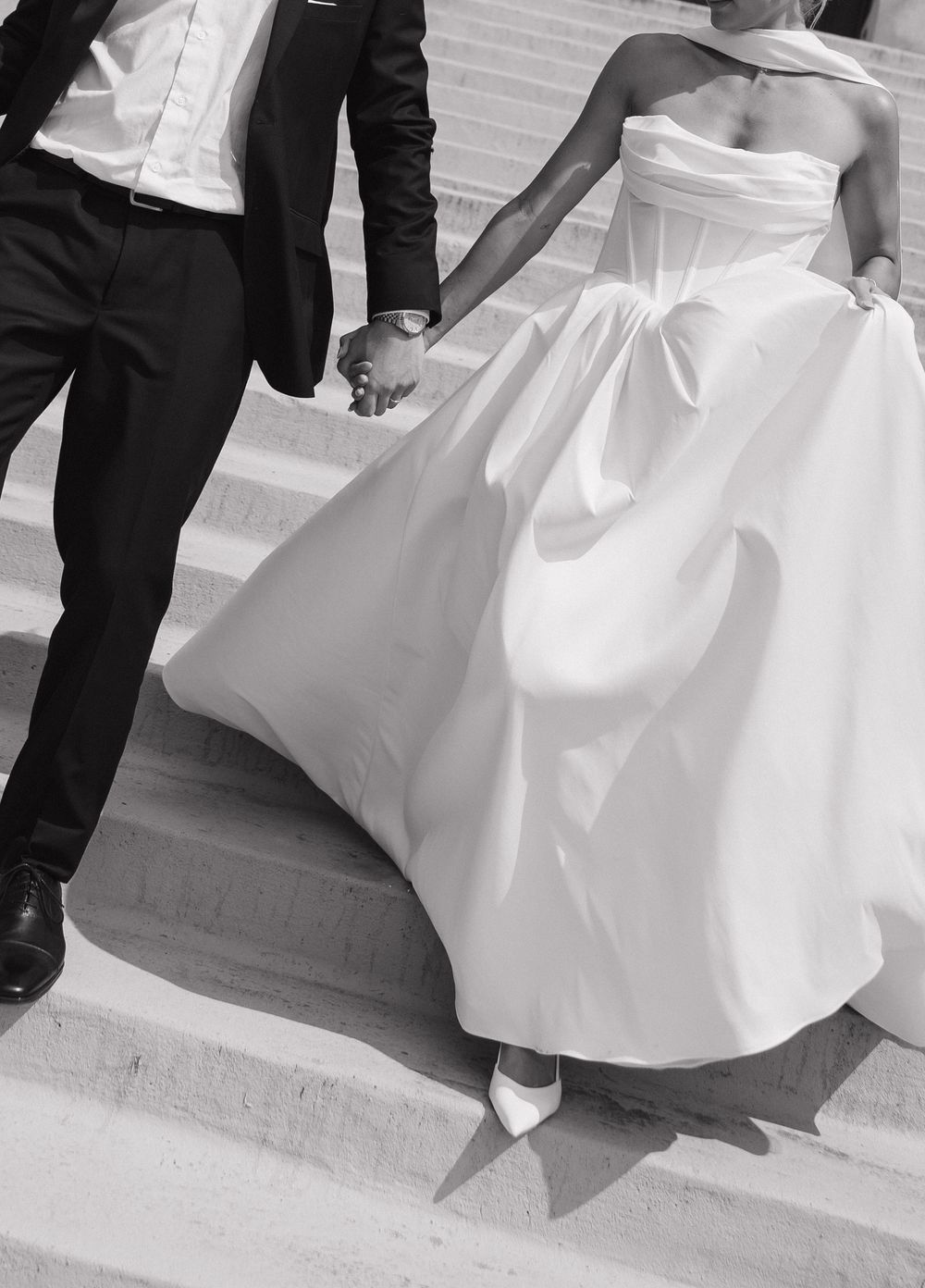 A couple in formal wedding attire walks down steps with the bride's white dress flowing behind them in black and white.