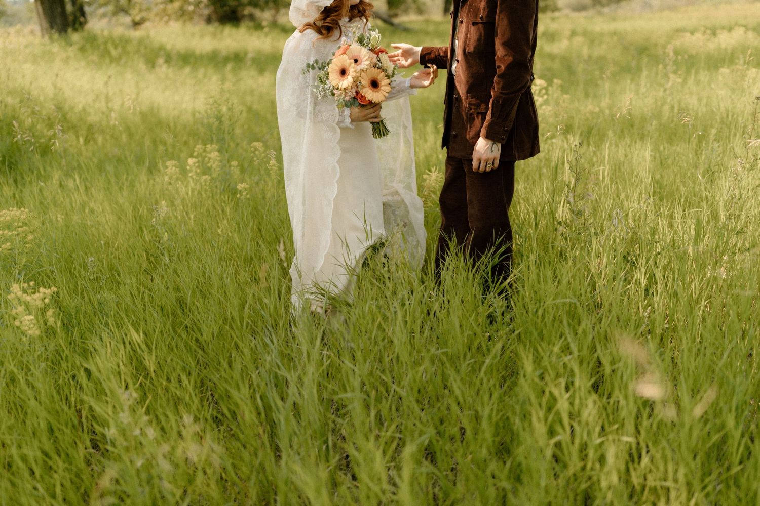 Wedding couple stands in tall summer grass holding orange and peach bouquet.