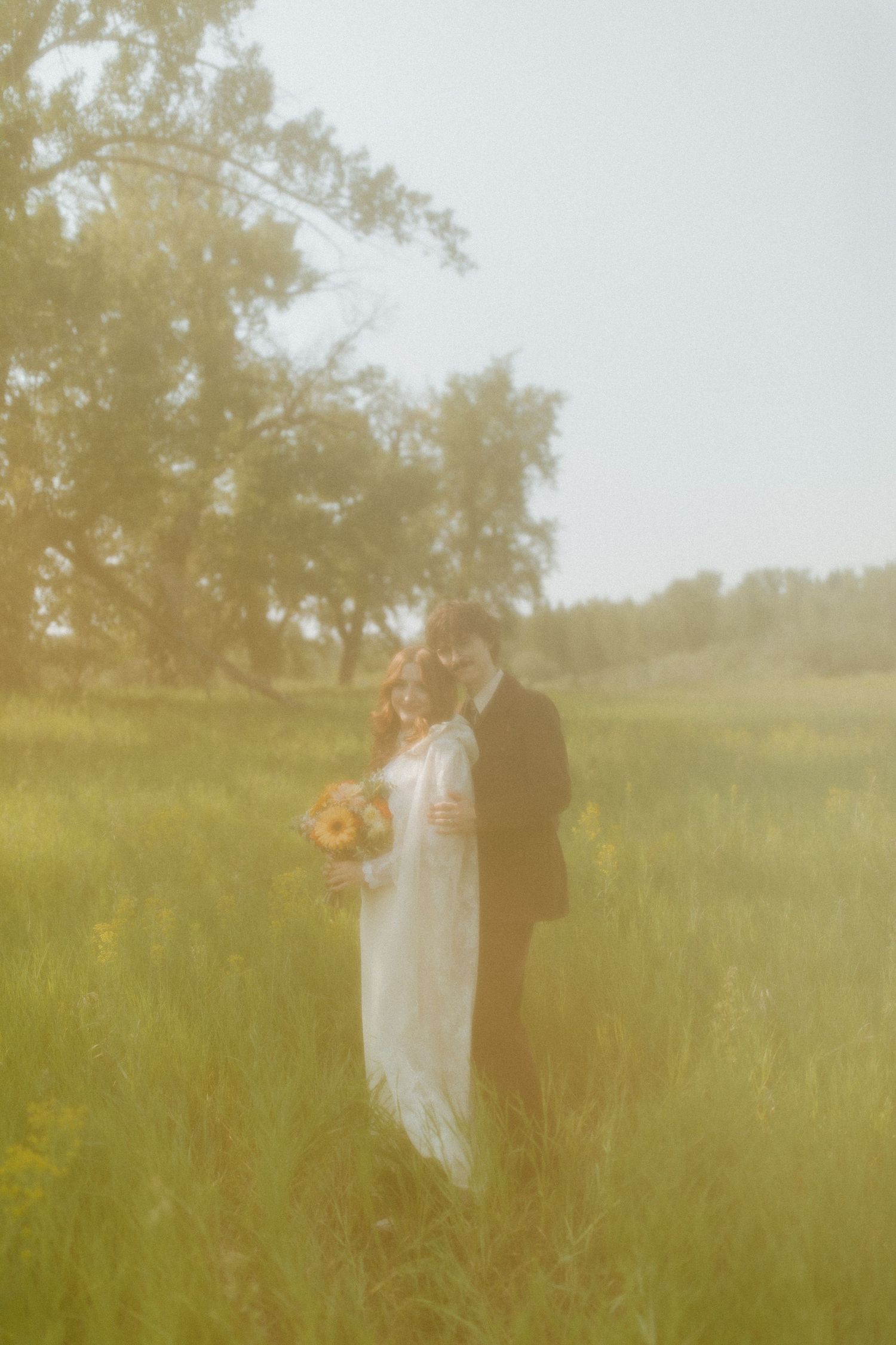 Vintage-style wedding photo with couple standing in sun-drenched meadow.