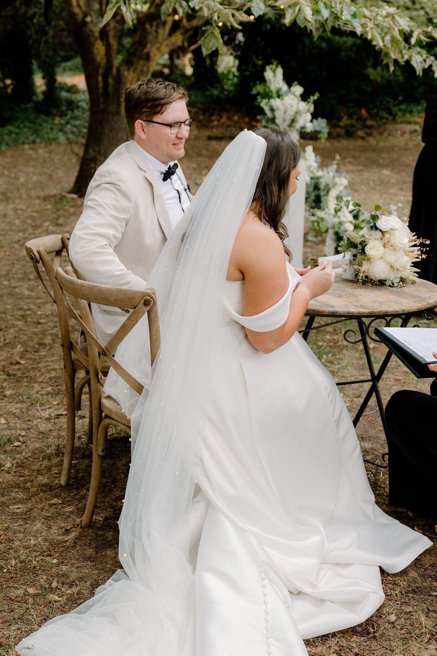 Bride and groom seated at outdoor wedding ceremony under trees with white floral decorations.