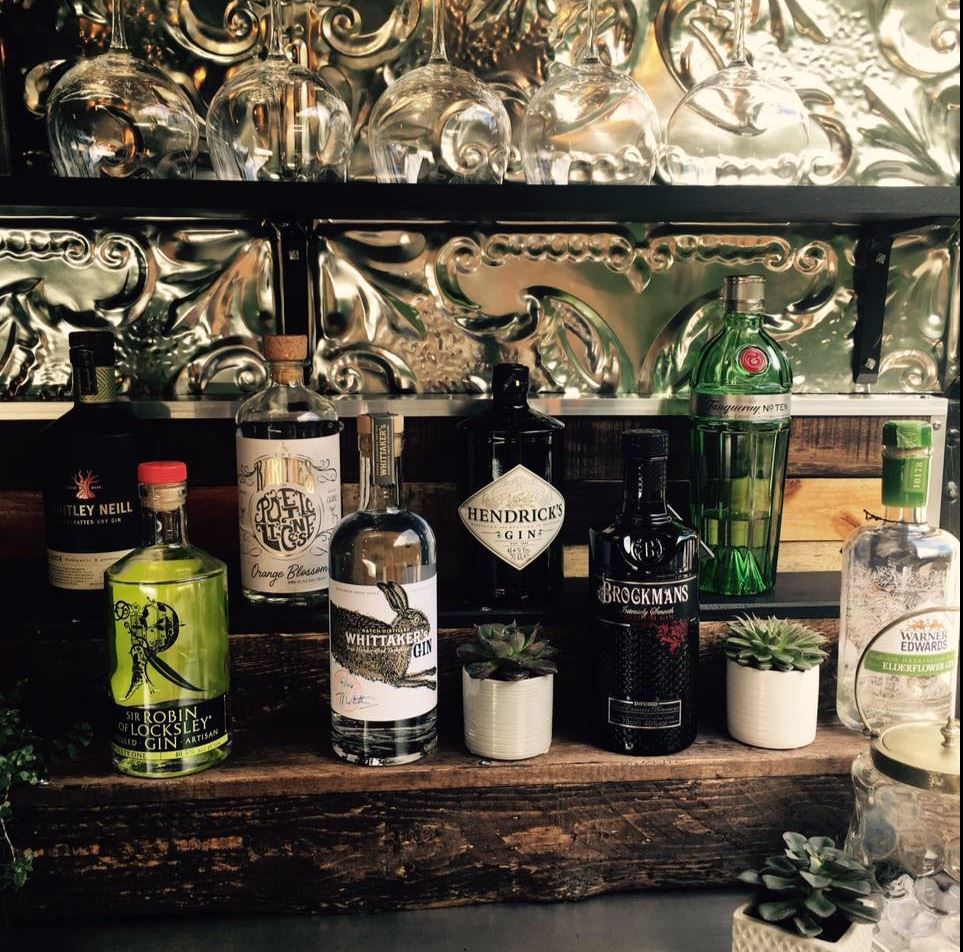 Various liquor bottles and craft cocktail ingredients arranged on a dark wooden shelf with mirrored backing.