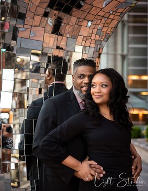 A couple embraces in front of a decorative mirrored mosaic wall, both wearing black and smiling warmly in an elegant setting during session.