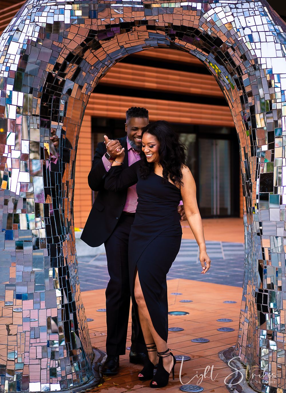 Engagement photoshoot of a couple in formal attire poses under a decorative mosaic mirror archway against an orange wall.