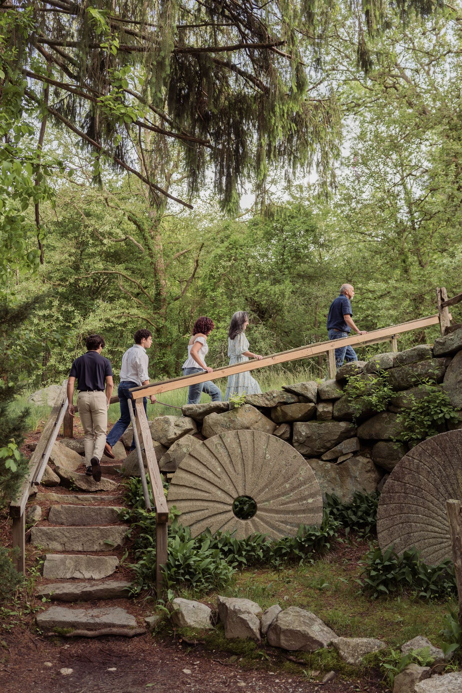 A group walks across a rustic stone bridge with old millstones displayed along a landscaped garden path.