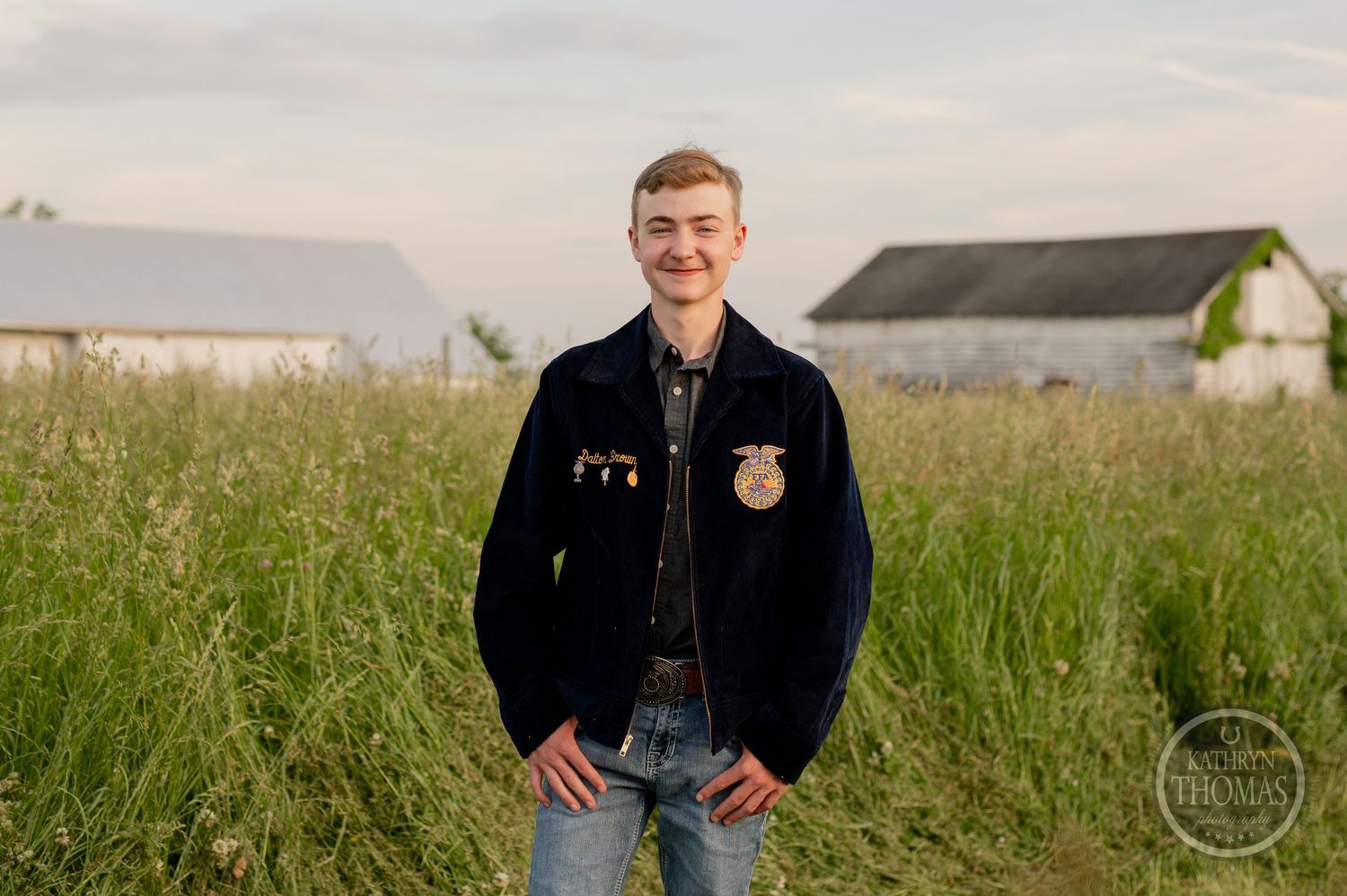 Student in FFA jacket stands smiling in field with farm buildings in background.