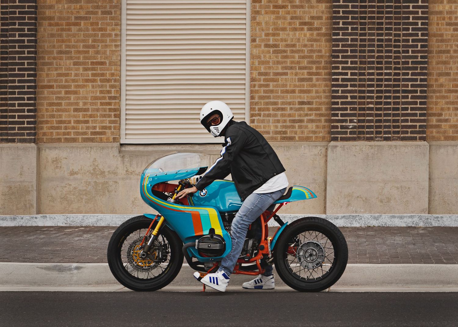 A motorcyclist in full gear seated on a turquoise vintage-style cafe racer motorcycle against a brick wall.