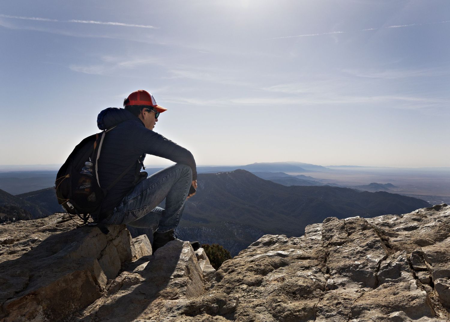 A hiker sitting on a mountain peak overlooking a vast landscape of rolling hills.