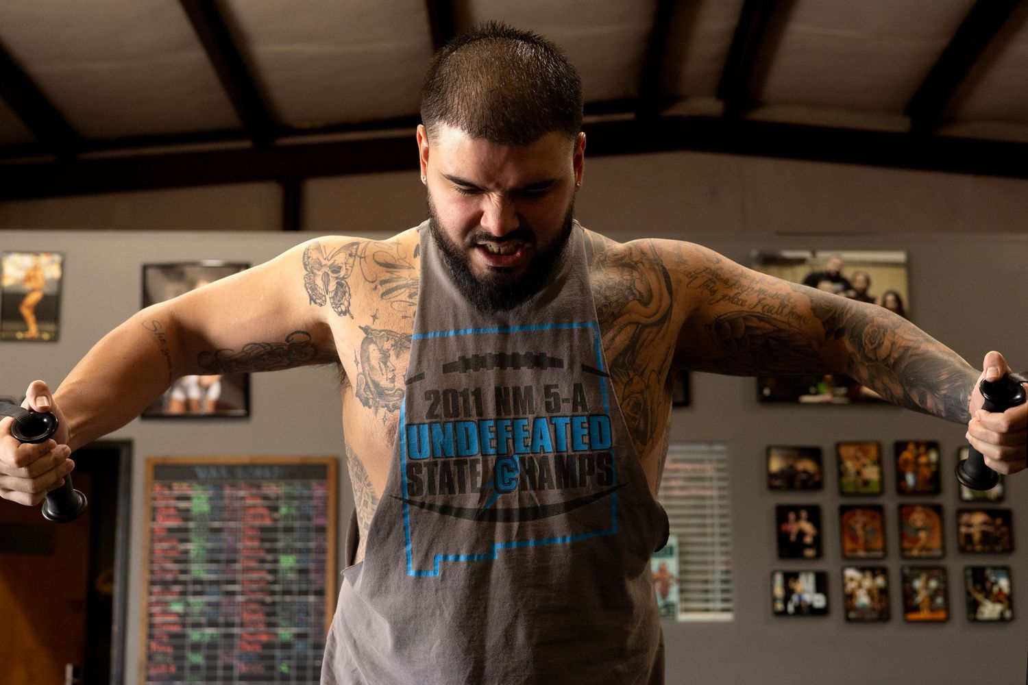 Strong athlete with tattoos stretches arms wide in gym setting.