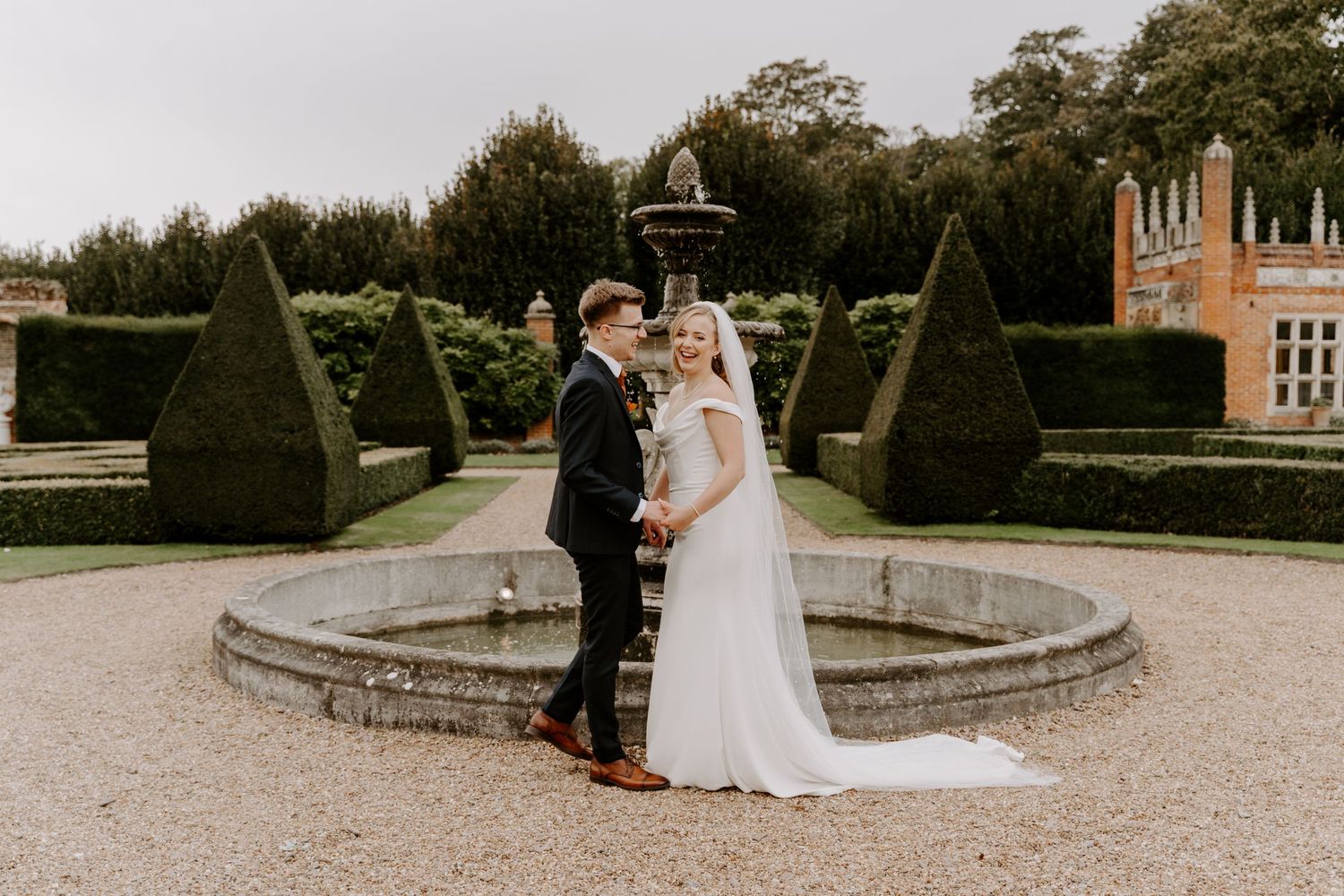 a bride and groom are stood in the grounds of oxnead hall. the bride is laughing at the camera and the groom is looking at his bride.