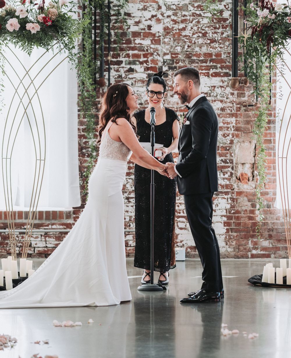 A wedding ceremony takes place under an arch with floral arrangements against a brick wall.
