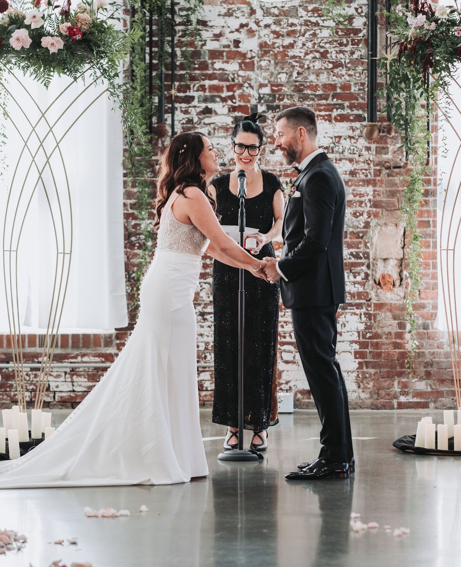 A wedding ceremony takes place under an arch with floral arrangements against a brick wall.