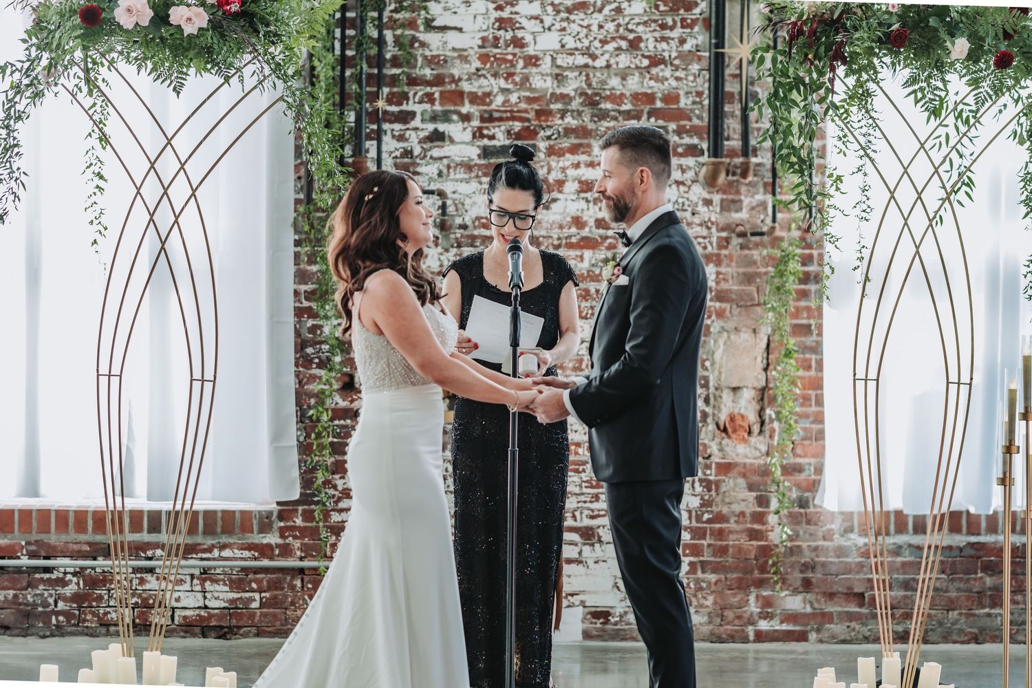 Wedding ceremony in progress under geometric arch with exposed brick backdrop.