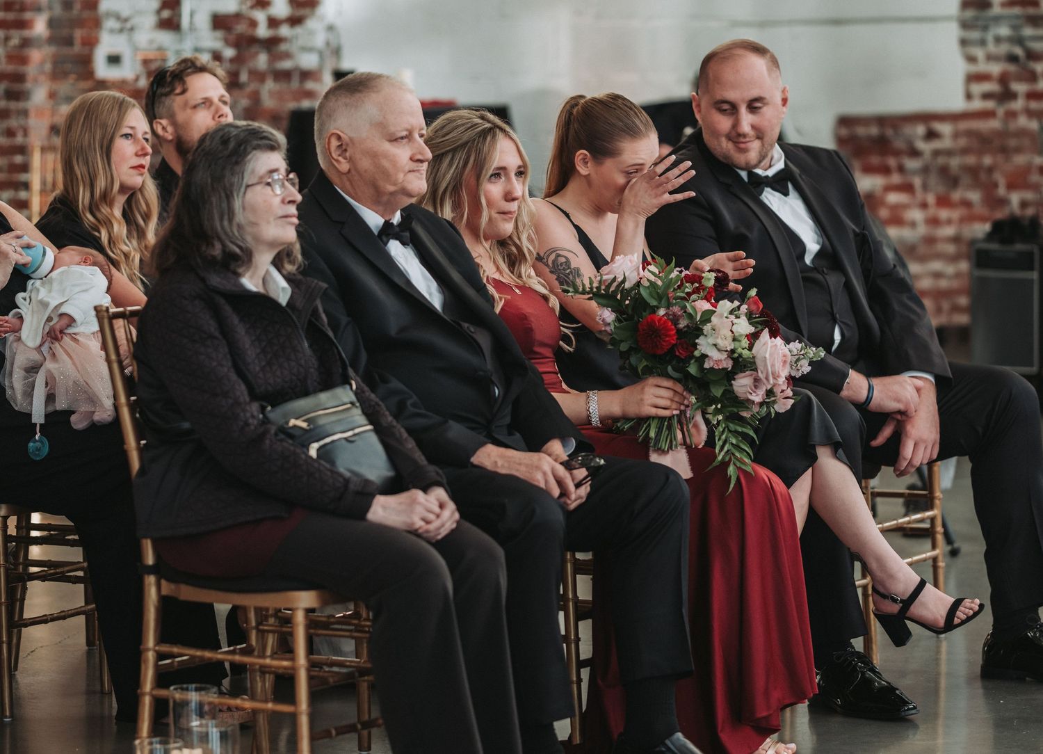 Wedding guests in formal attire seated on wooden chairs in a brick-walled venue.