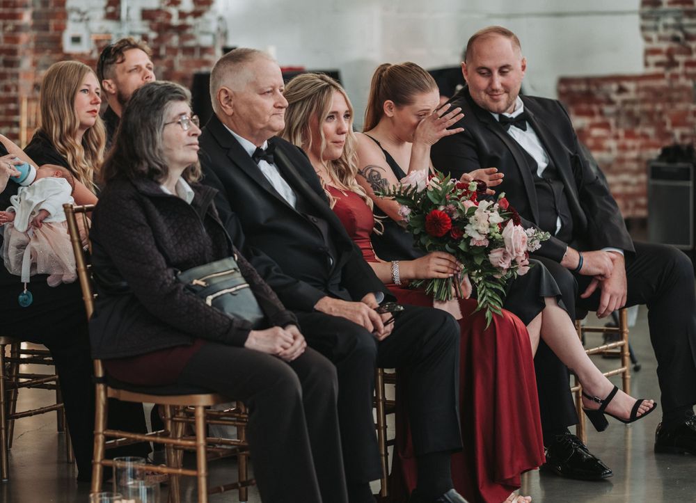 Wedding guests in formal attire seated on wooden chairs in a brick-walled venue.