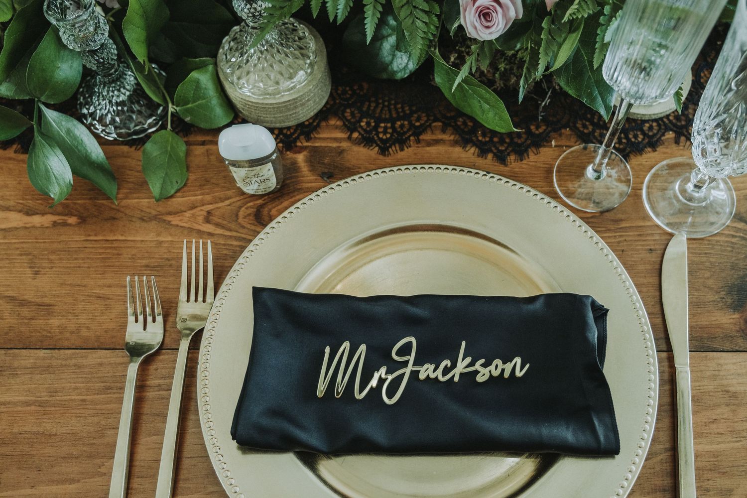 Elegant place setting with black napkin personalized with 'Mr Jackson' text and floral decor on wooden table.