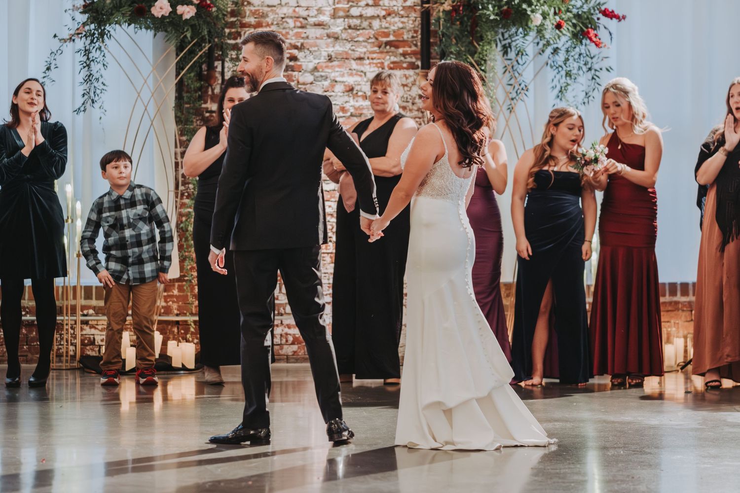Wedding couple dancing with guests in elegant attire at a rustic brick venue.