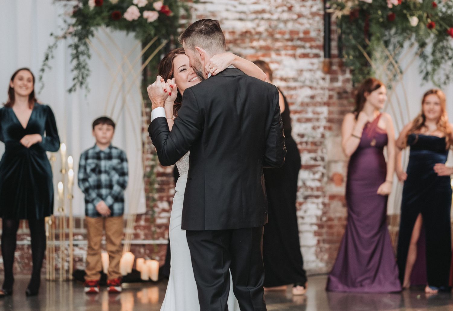 A couple shares their first dance at a wedding reception in front of an exposed brick wall decorated with greenery.