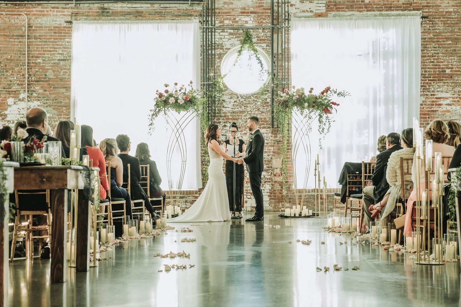 Indoor wedding ceremony with white draping, floral arch, and exposed brick walls.