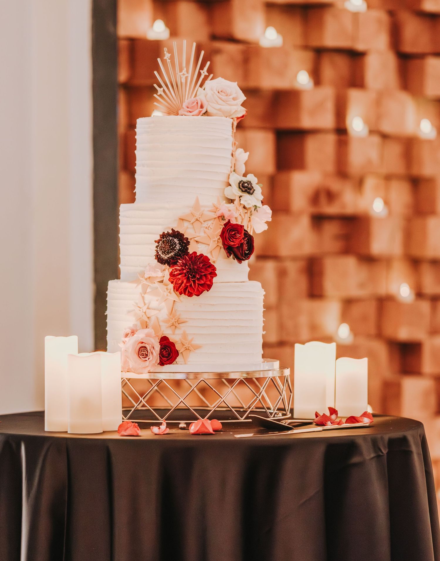 Elegant white wedding cake with floral decorations and candles against textured wall.