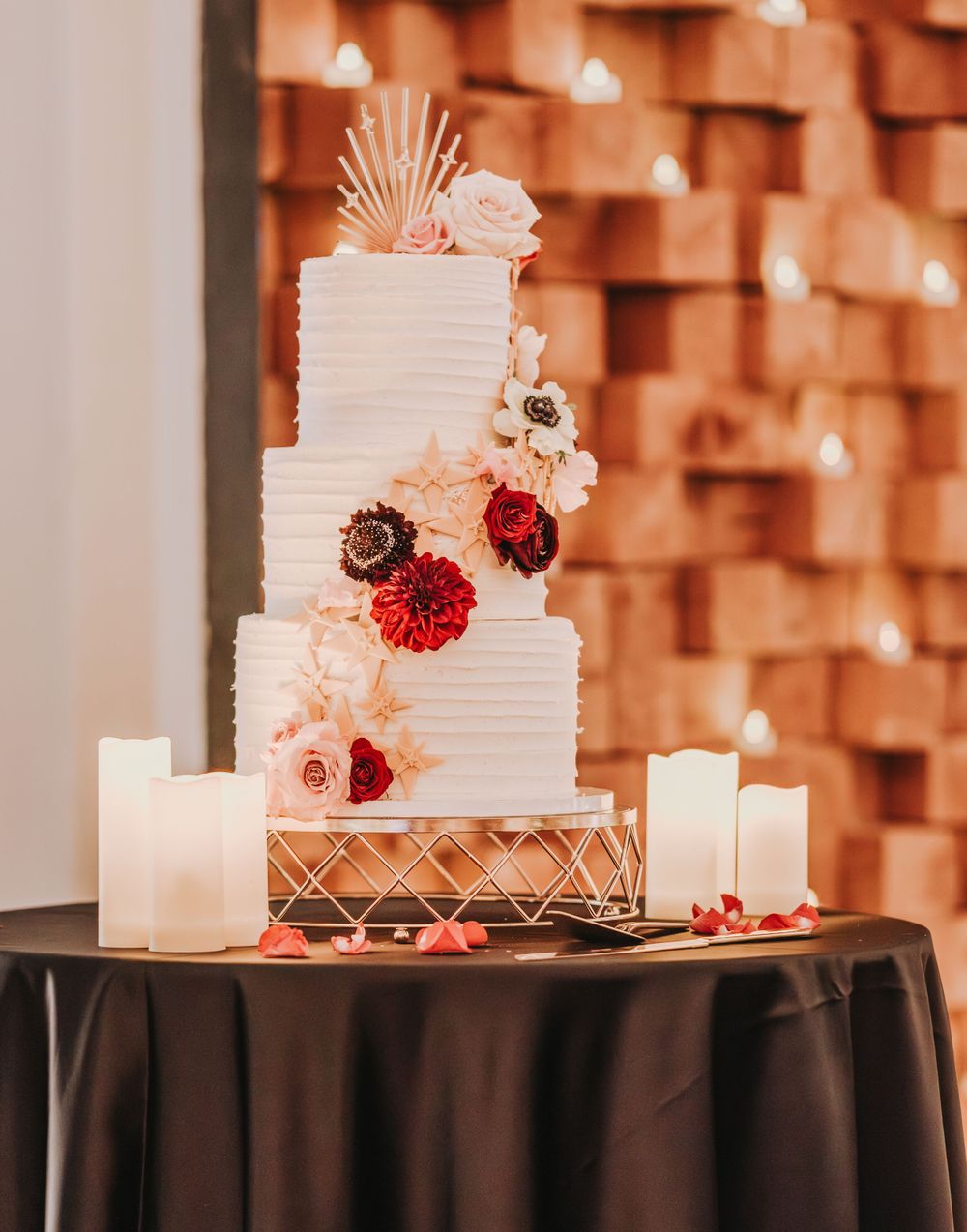 Elegant white wedding cake with floral decorations and candles against textured wall.