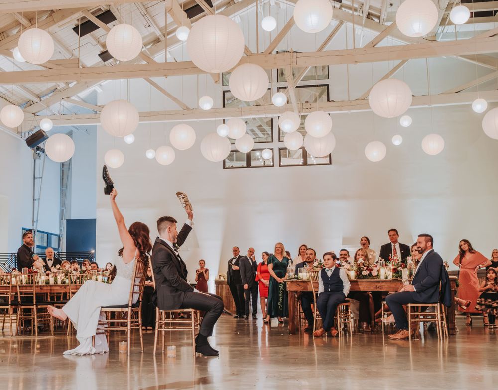 Wedding guests cheer as couple performs energetic dance moves in reception venue with hanging lanterns.
