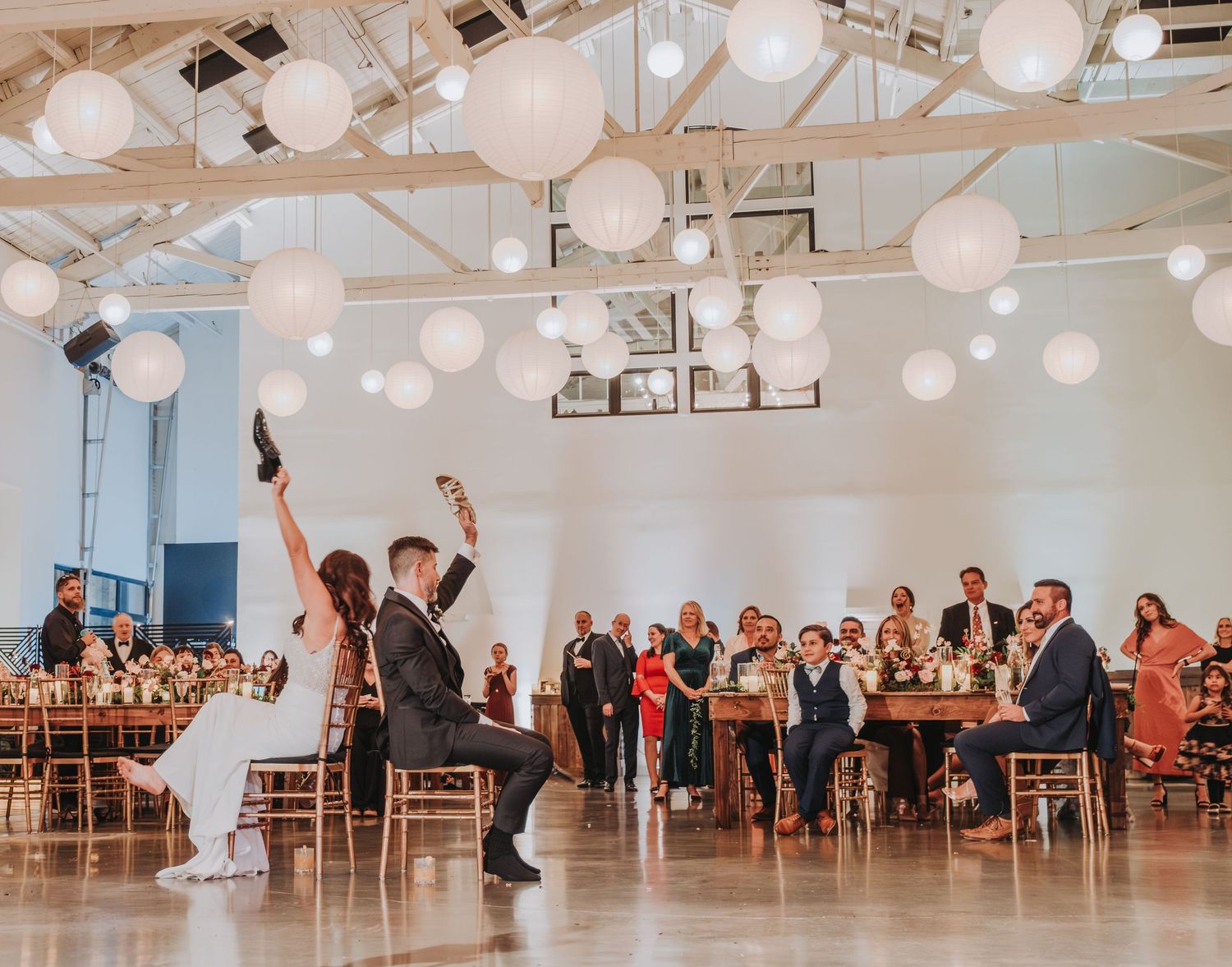 Wedding guests cheer as couple performs energetic dance moves in reception venue with hanging lanterns.