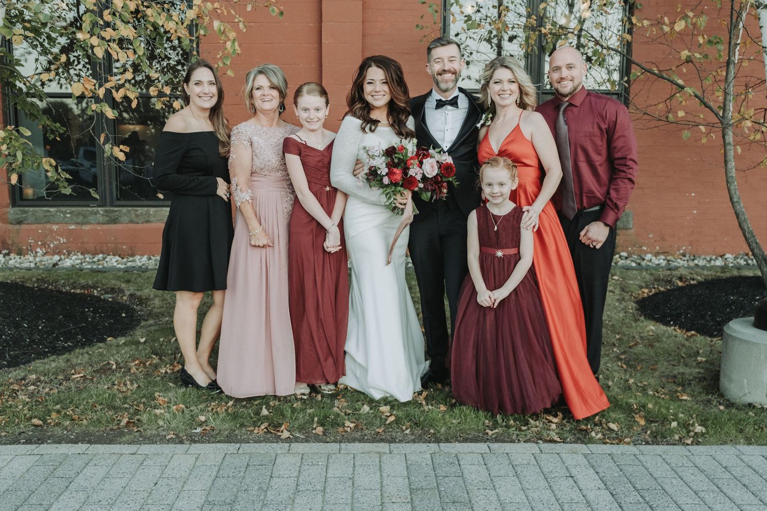 Wedding party in autumn colors poses outdoors with bridesmaids wearing burgundy, coral and blush dresses against brick wall.