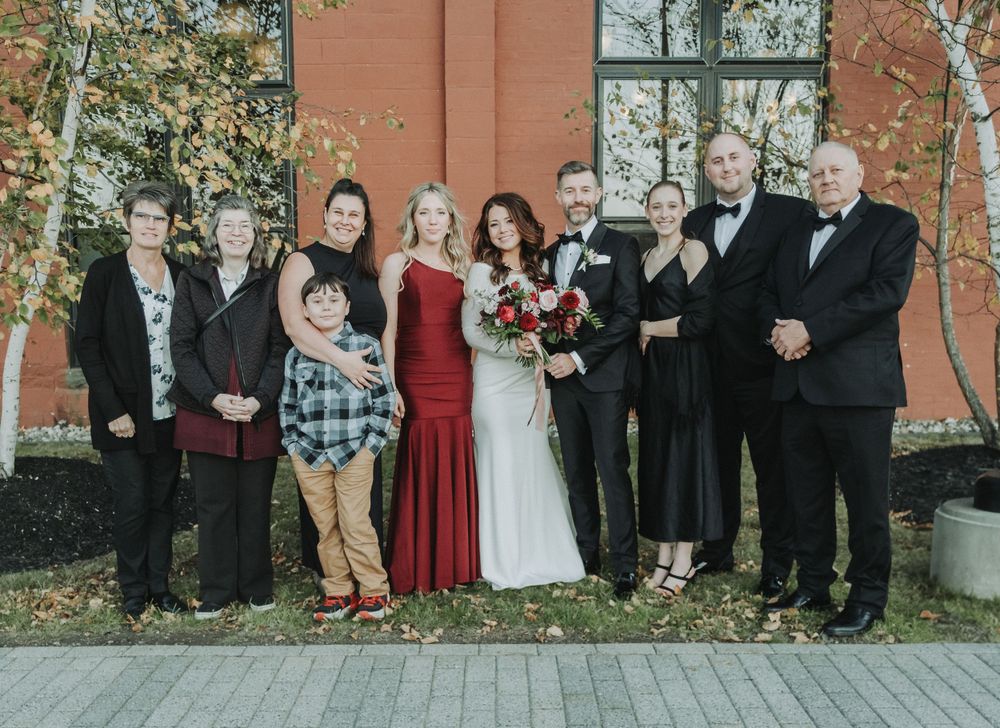 A family group poses outdoors against a brick wall wearing formal attire at a wedding celebration.