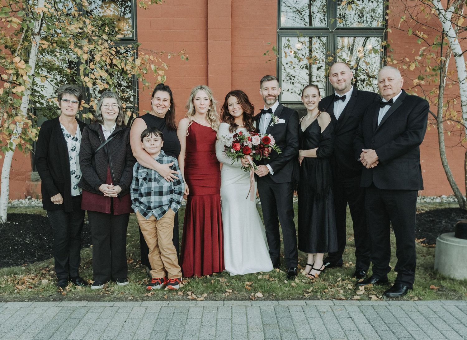 A family group poses outdoors against a brick wall wearing formal attire at a wedding celebration.