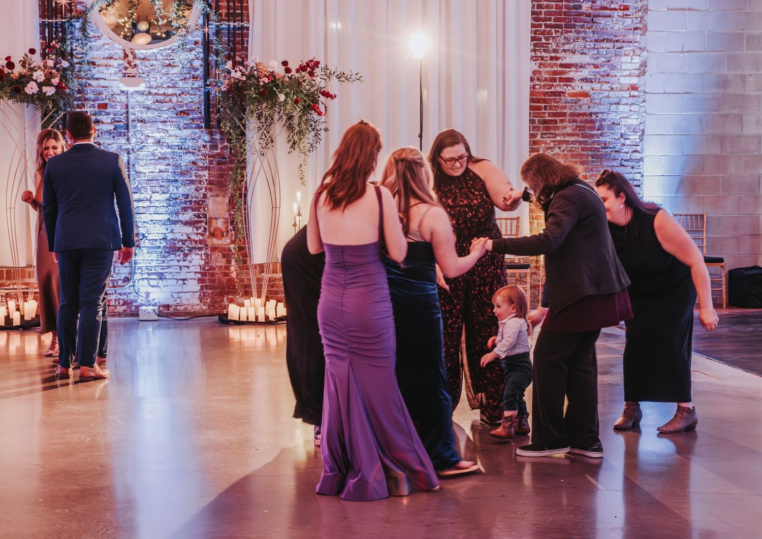 Wedding guests dance together in elegant reception venue with exposed brick walls and mood lighting.