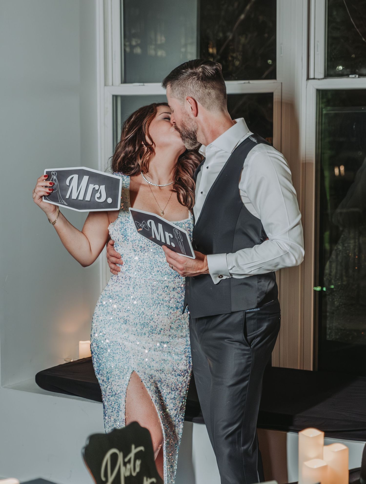 Newlyweds share a romantic kiss while holding Mr and Mrs signs at their wedding reception.