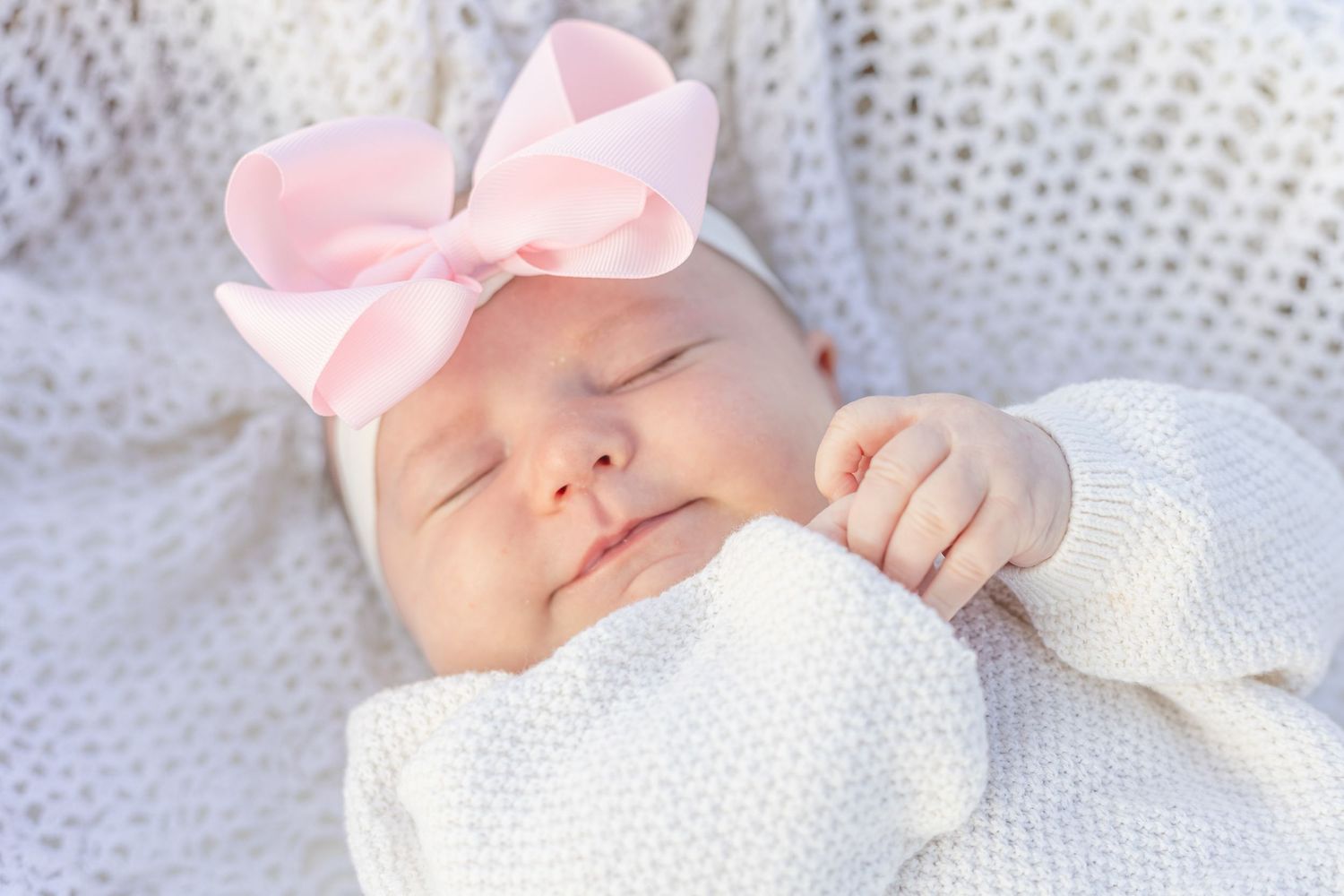 Peaceful newborn baby sleeping with pink bow headband, wrapped in soft white knit blanket.