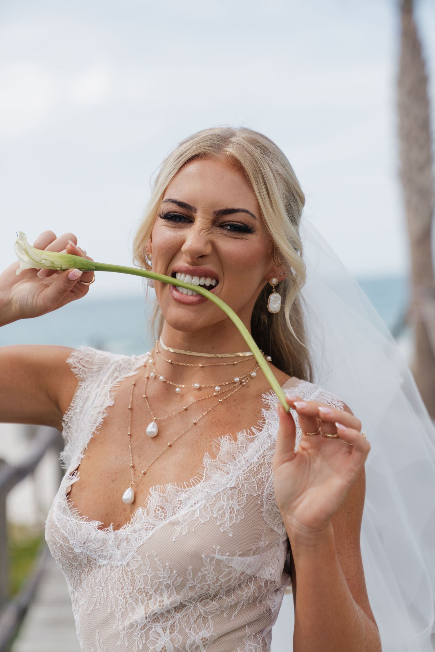 Blonde bride in lace wedding dress and veil playfully biting a green stem outdoors near the ocean.