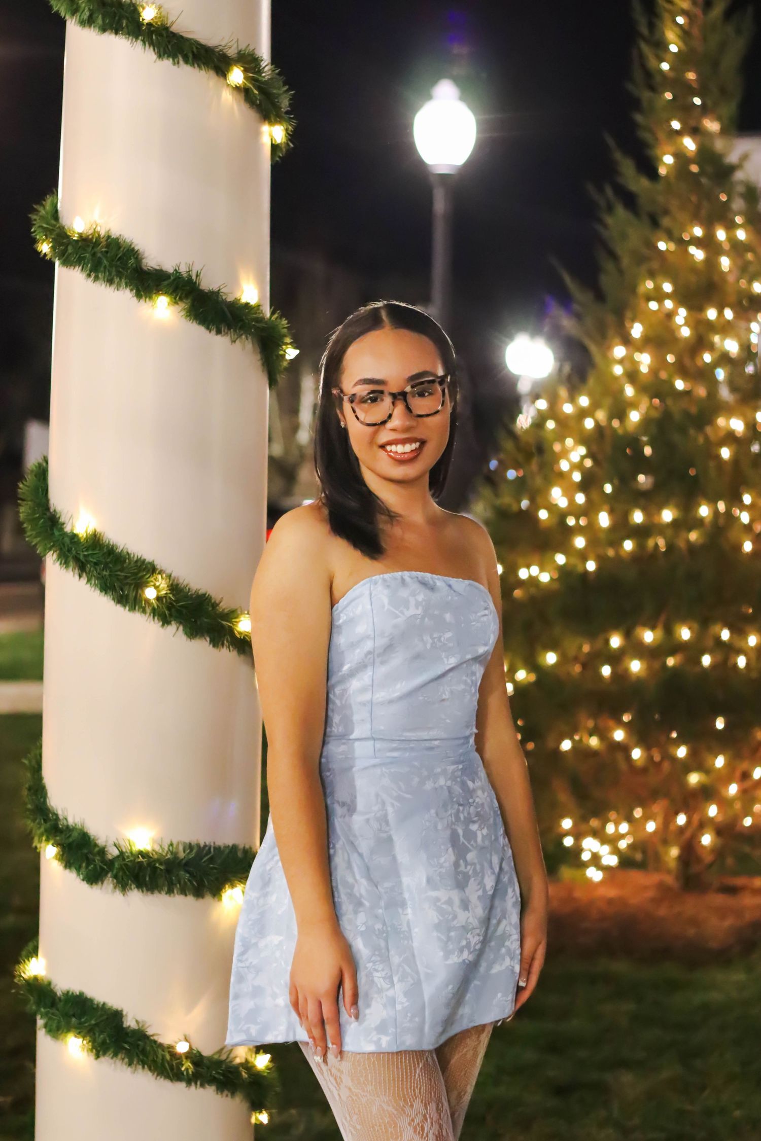 Woman in light blue dress stands by holiday decorations and lit Christmas trees.