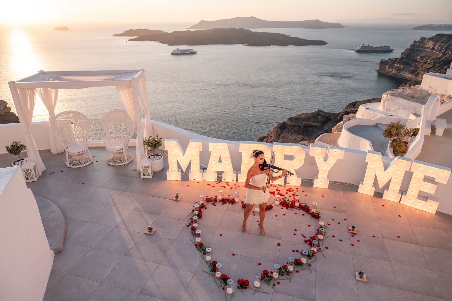 Dreamy proposal setup with lit 'Marry Me' sign and white gazebo overlooking Santorini.