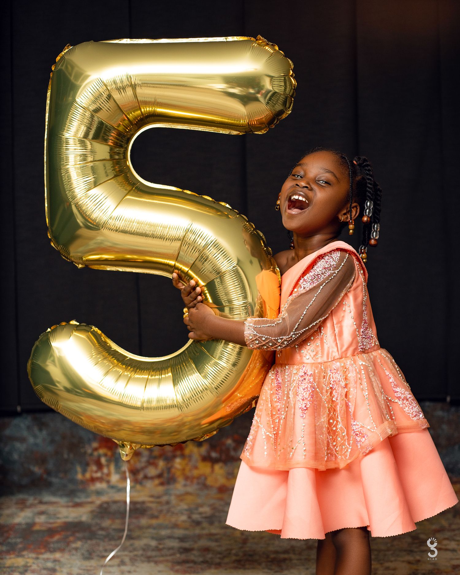 Young child in peach dress celebrating with large gold number 5 balloon.