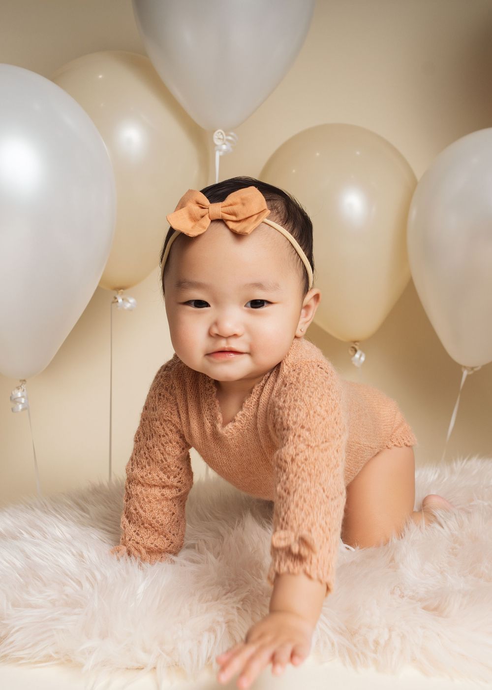 Baby girl in tan lace romper and bow headband crawling on fur rug surrounded by cream balloons.