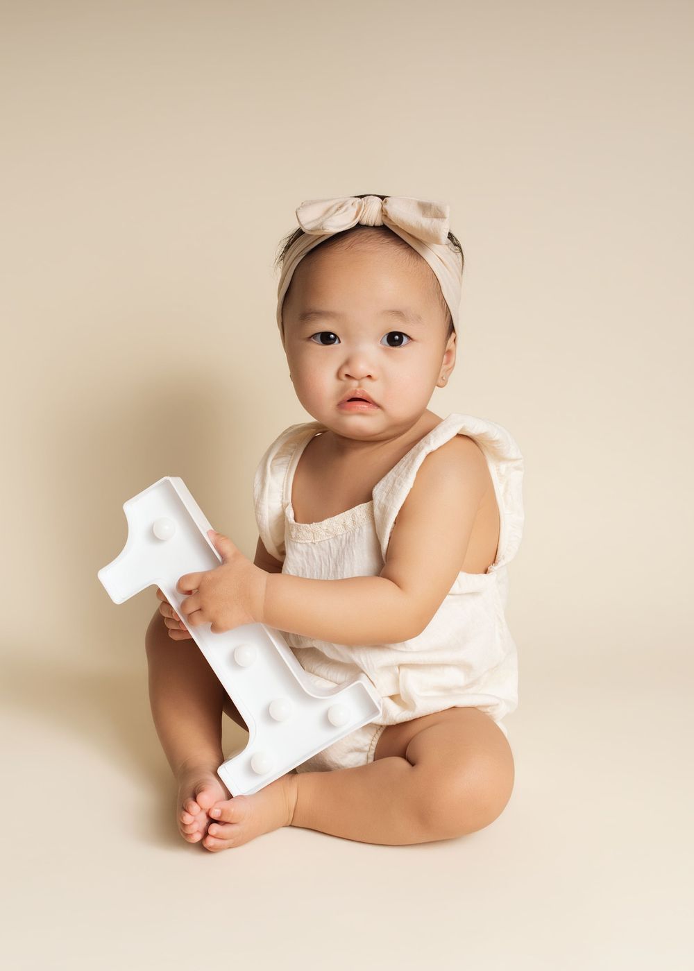 Baby girl in cream romper and bow headband holds a number 1 sign, sitting for a first birthday portrait.