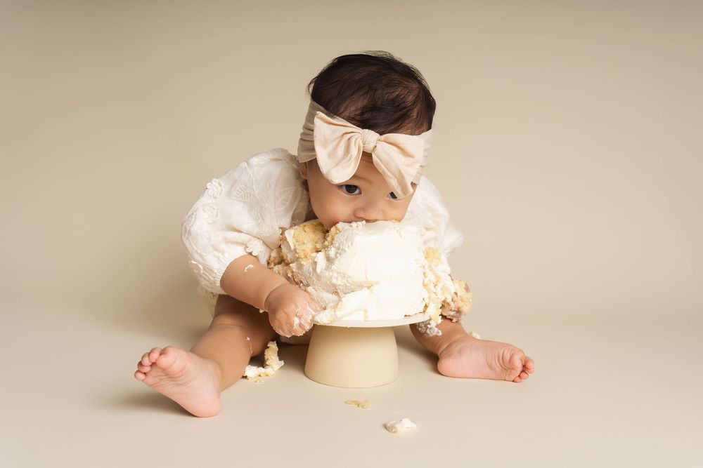 Baby girl smashes white first birthday cake wearing a white romper and pink bow headband.
