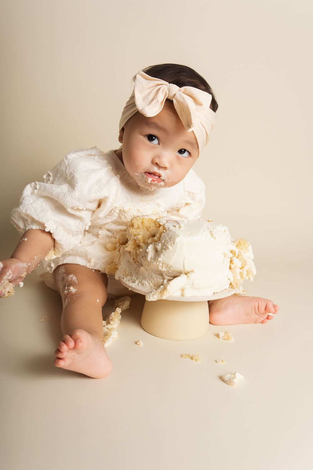 Baby girl in white dress with bow headband smashes first birthday cake during milestone smash cake photo session.