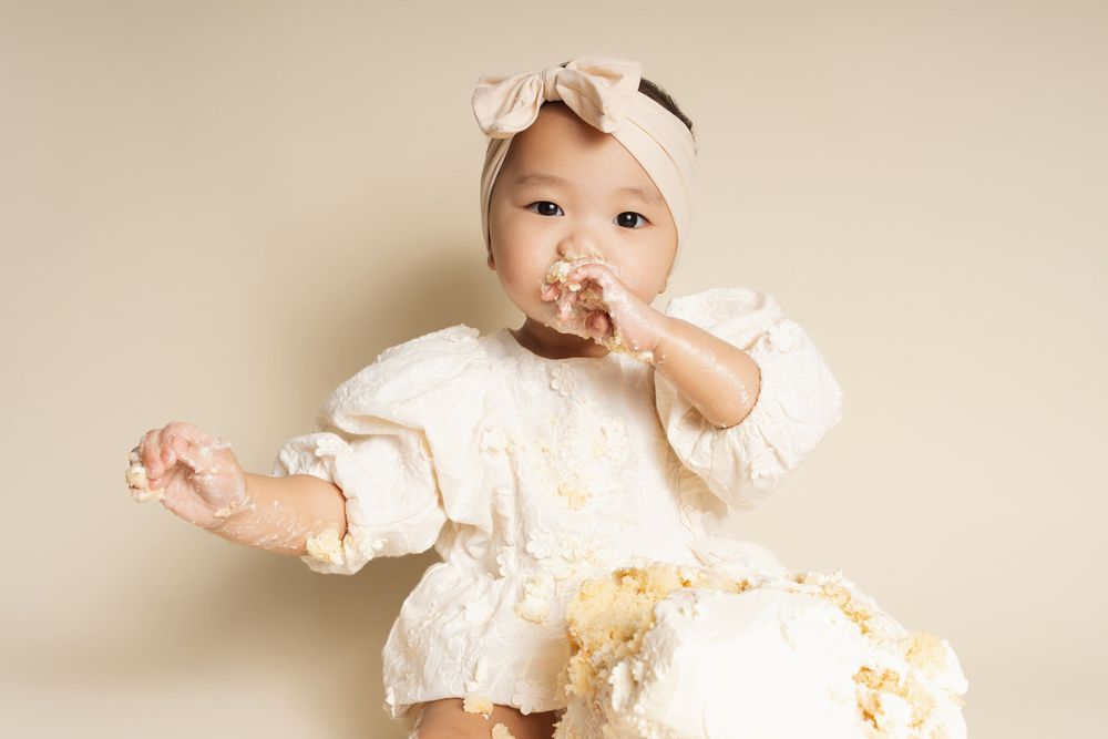 Baby girl in white dress and bow headband eating smash cake during first birthday photo session.