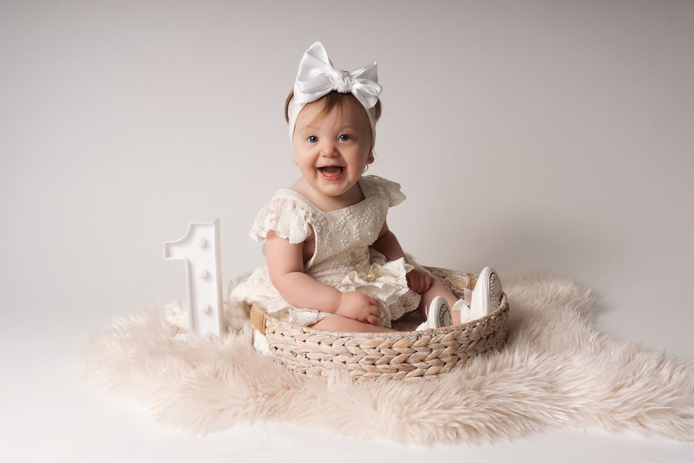 A baby wearing a white bow sits on a fluffy cream-colored rug during a milestone photo session.