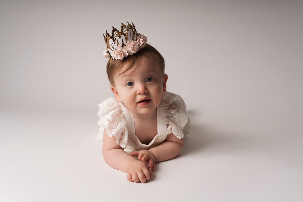 Baby wearing pink crown and white ruffled dress lies on light backdrop during professional photo session.