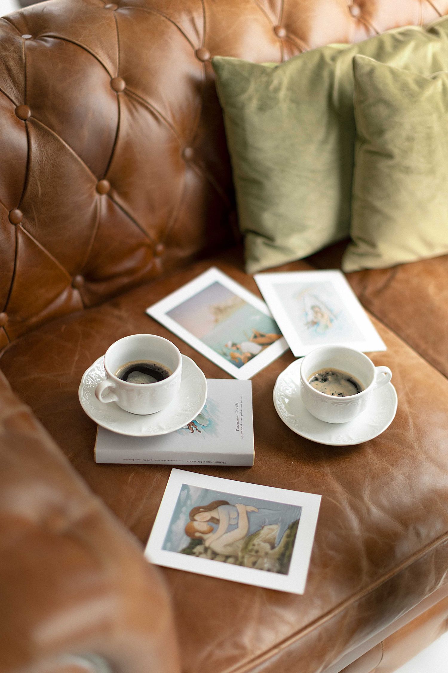 Two coffee cups and photographs arranged on a brown leather couch with green accent pillows in a casual setting.