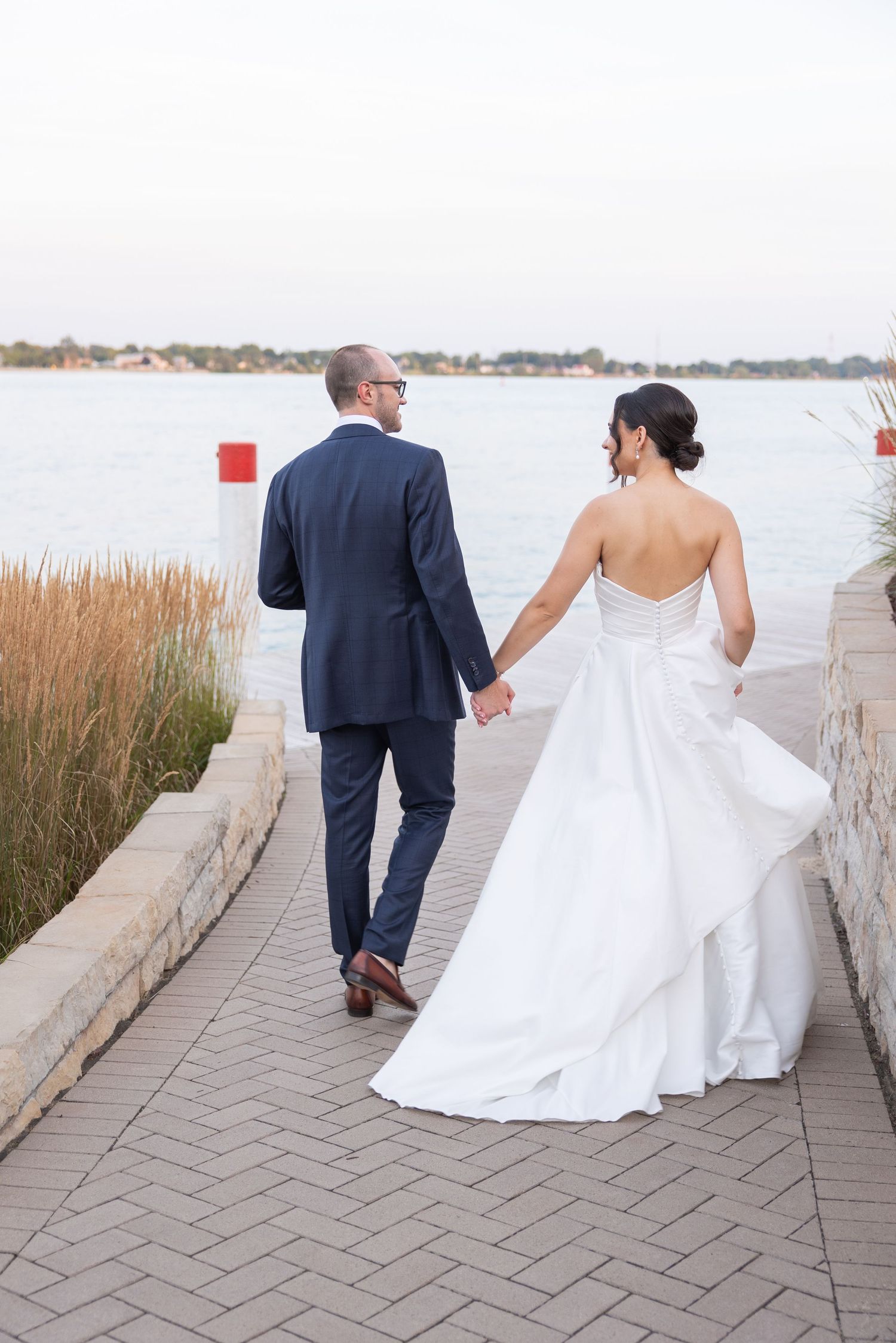 A couple in wedding attire walks hand in hand along a waterfront path bordered by tall grass at sunset. Metro Detroit Wedding Southeast MI