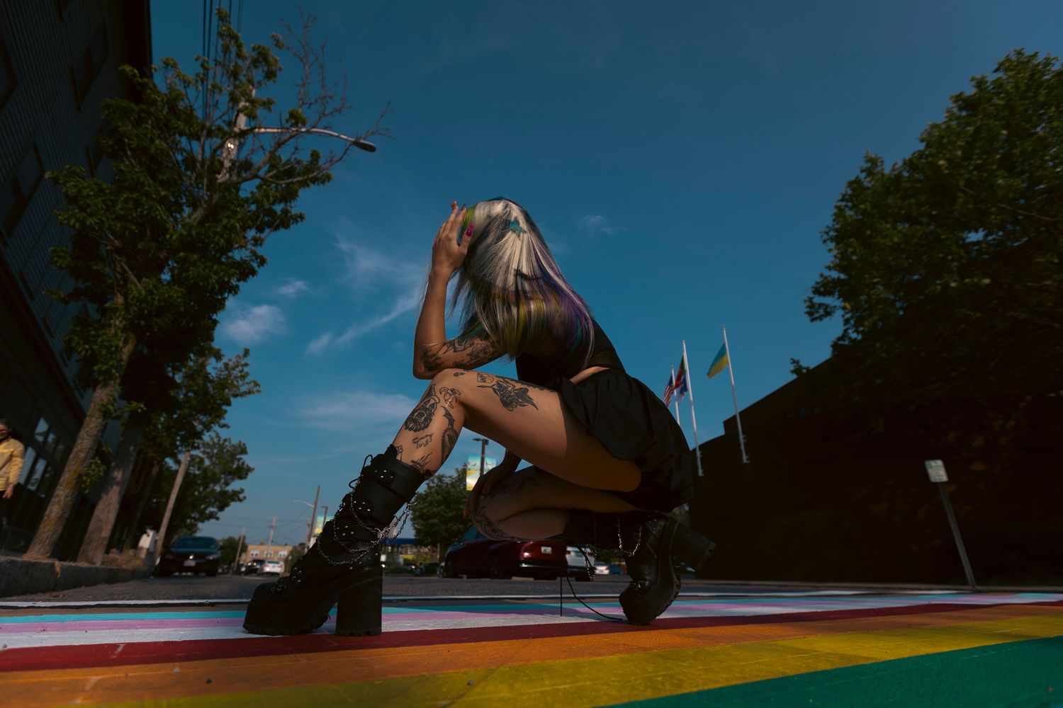 Person in black outfit and platform boots crouching on a colorful rainbow street surface during a sunny day.