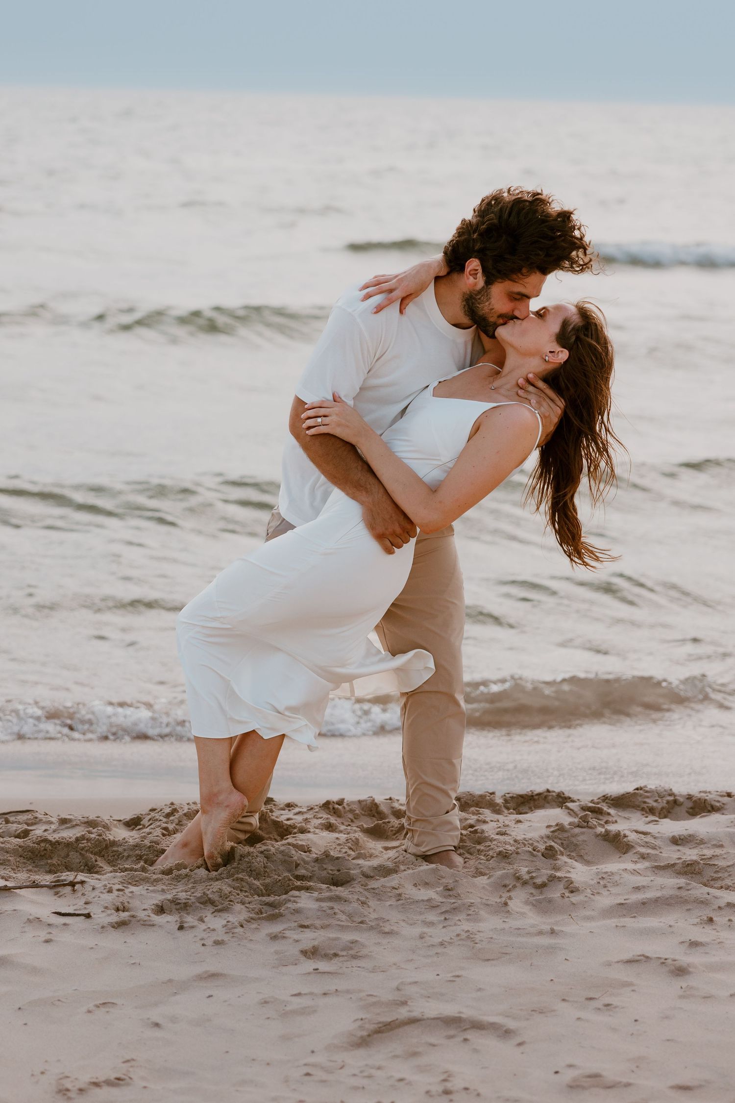 A romantic couple in white outfits dances on the beach as they dip, twirl and embrace during their seaside photoshoot. Detroit Wedding Photo